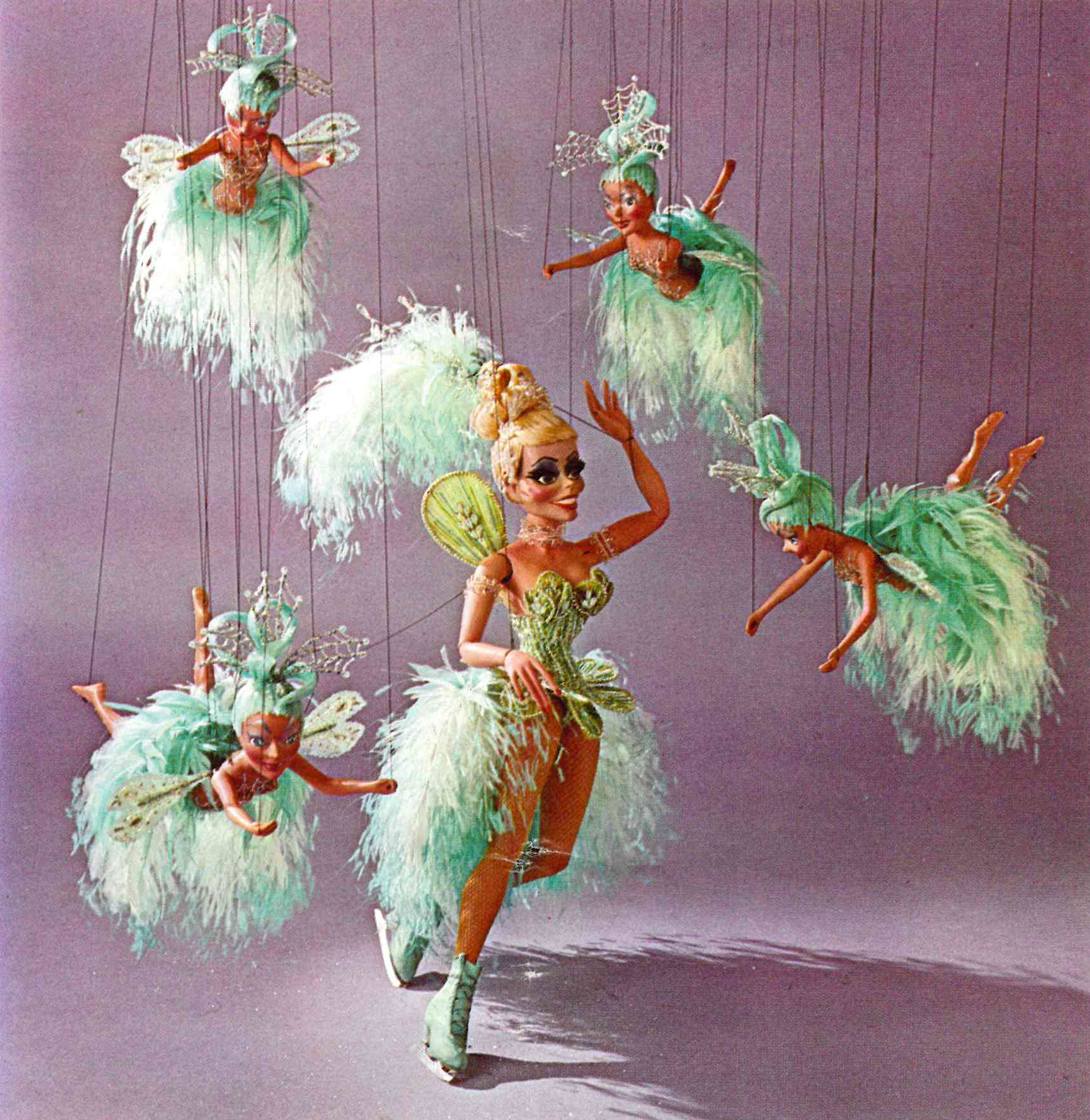 Color photograph of five tanned fairy puppets in green feathered outfits, one ice skating, the rest flying in the air